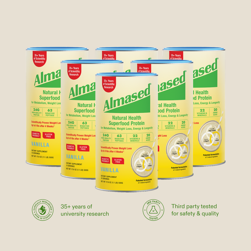 Five cans of Almased Natural Health Superfood Protein in vanilla flavor, arranged in a fan shape on a light beige background with two green certification logos at the bottom.